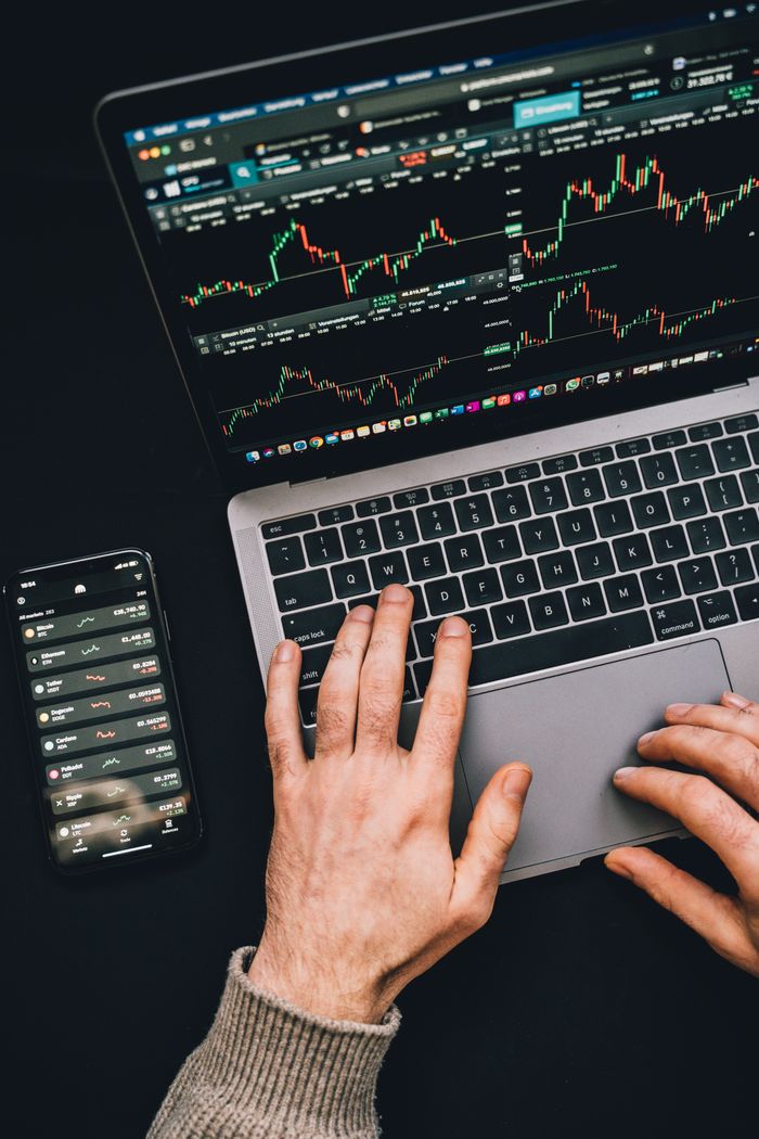 Hands typing on a laptop while cryptocurrency charts and a smartphone screen are visible, highlighting digital finance.